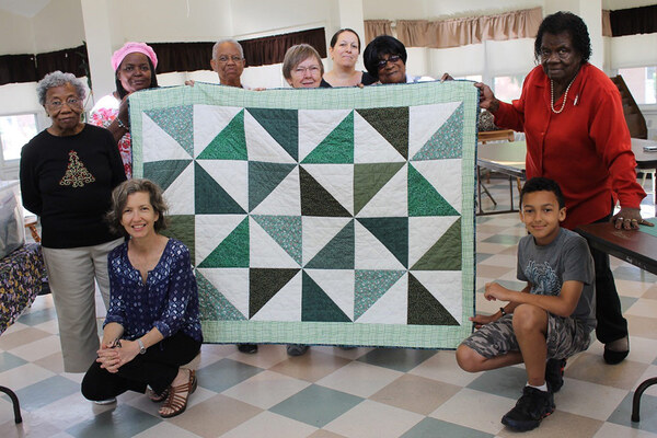 Seniors and children standing around and holding a completed quilt in a room